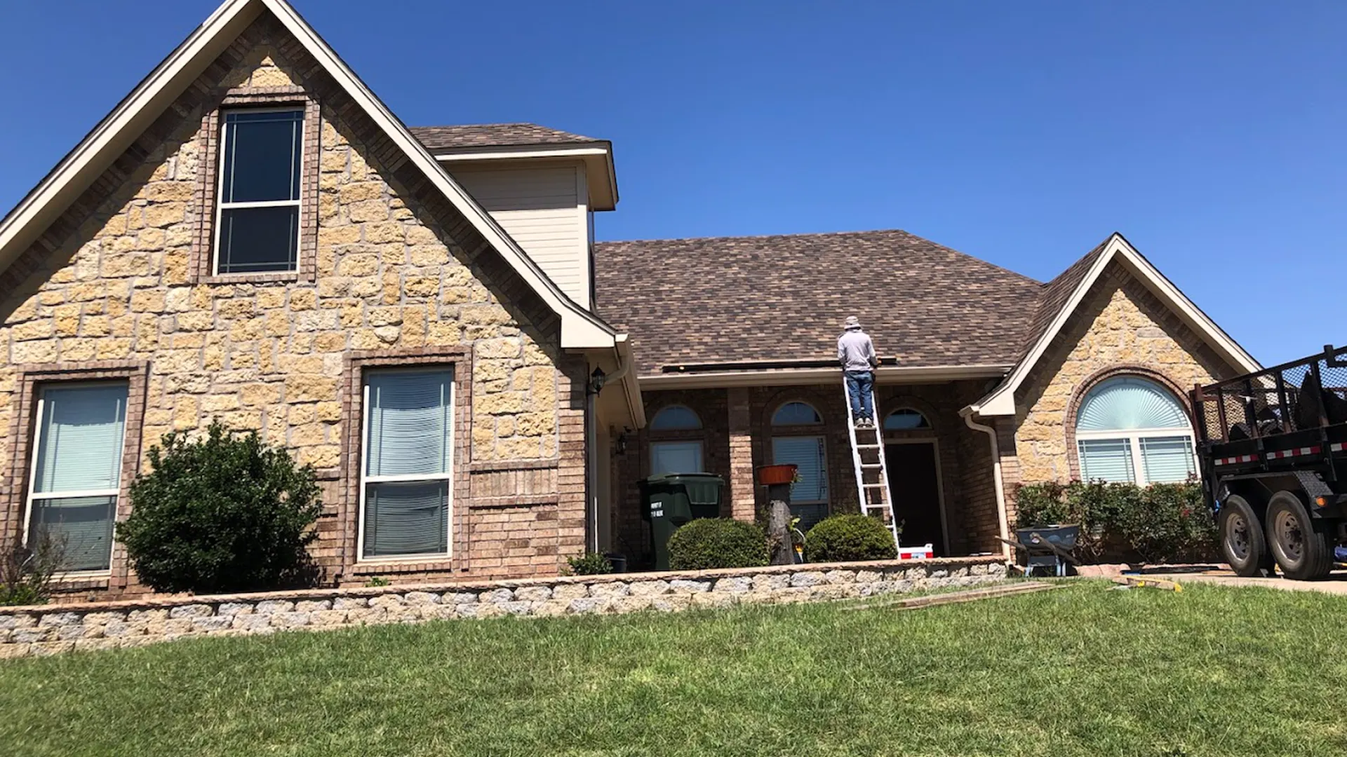Stone house with a ladder against the porch roof, a roofer up top and a work truck parked beside it